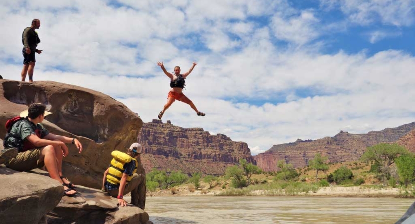 A person is suspended mid-air after jumping off a cliff into a river. Others look on, and everyone is wearing life jackets. Behind them all are tall, red canyon walls. 